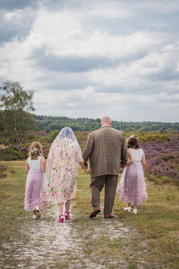 Catherine with her husband and twin daughters walking away from the camera in the New Forest. They are all in wedding attire.